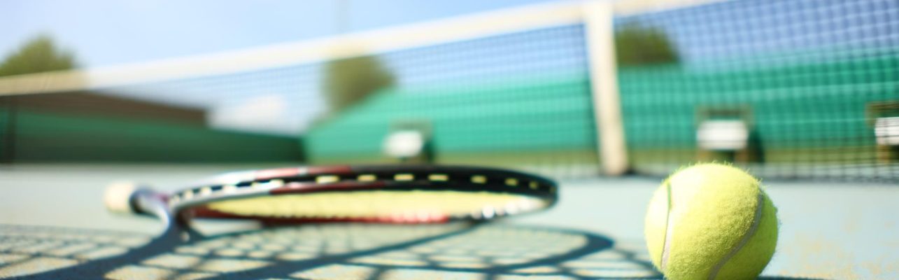 Une balle et une raquette de tennis sur un terrain ensoleillé avec le filet et des sièges flous en arrière-plan.