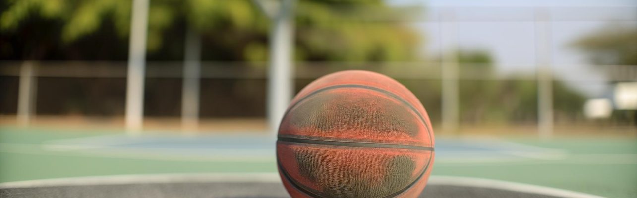 Un ballon de basket bien utilise repose sur un terrain exterieur avec un panier et une cloture en arriere-plan.