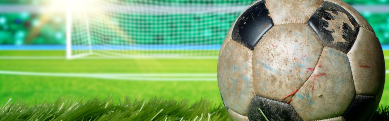 Un ballon de football usé posé sur de l'herbe verte avec un fond flou de terrain et de buts.