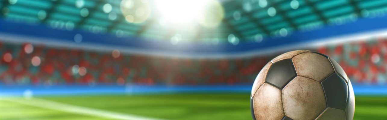 Un ballon de football sur l'herbe fraichement coupee devant les gradins flous d'un stade.
