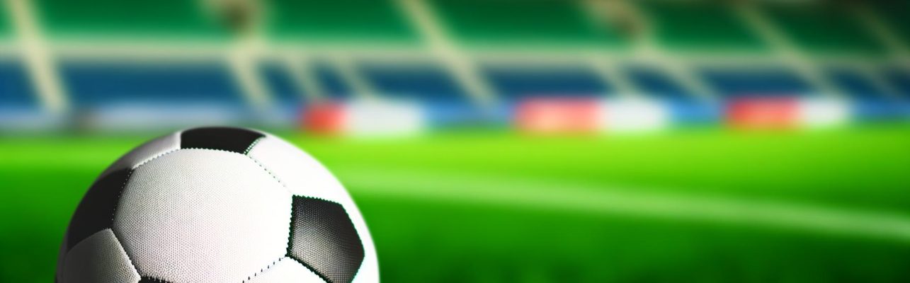 Un ballon de football pose sur un terrain vert avec un stade flou au loin.