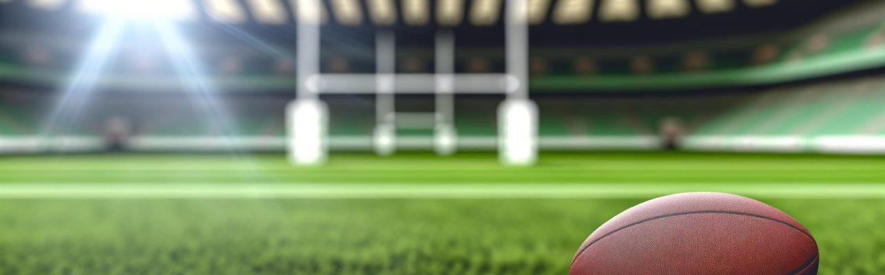 Un ballon de rugby repose sur l'herbe au centre du terrain sous la lumière naturelle du soleil, avec des gradins et des poteaux de but flous en arrière-plan.