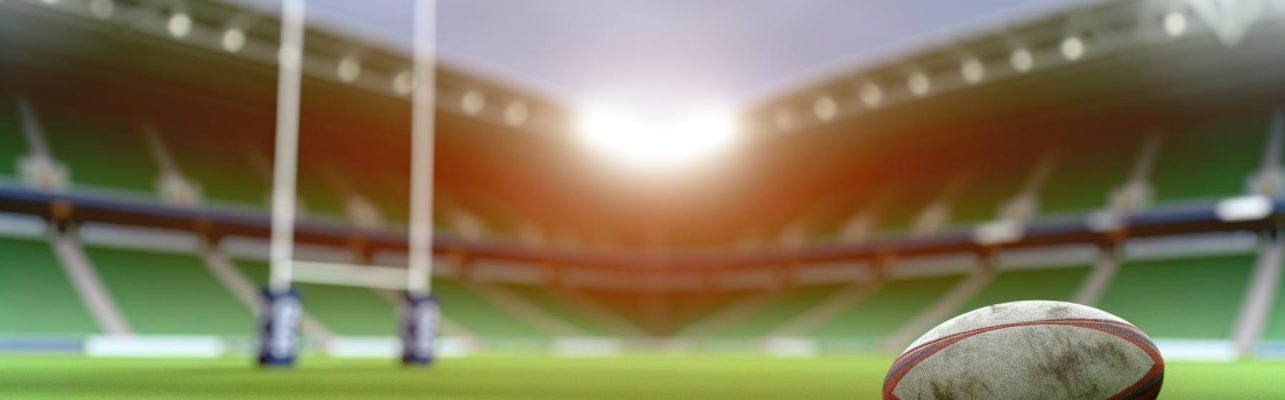 Un ballon de rugby repose sur un terrain avec des gradins en arrière-plan.