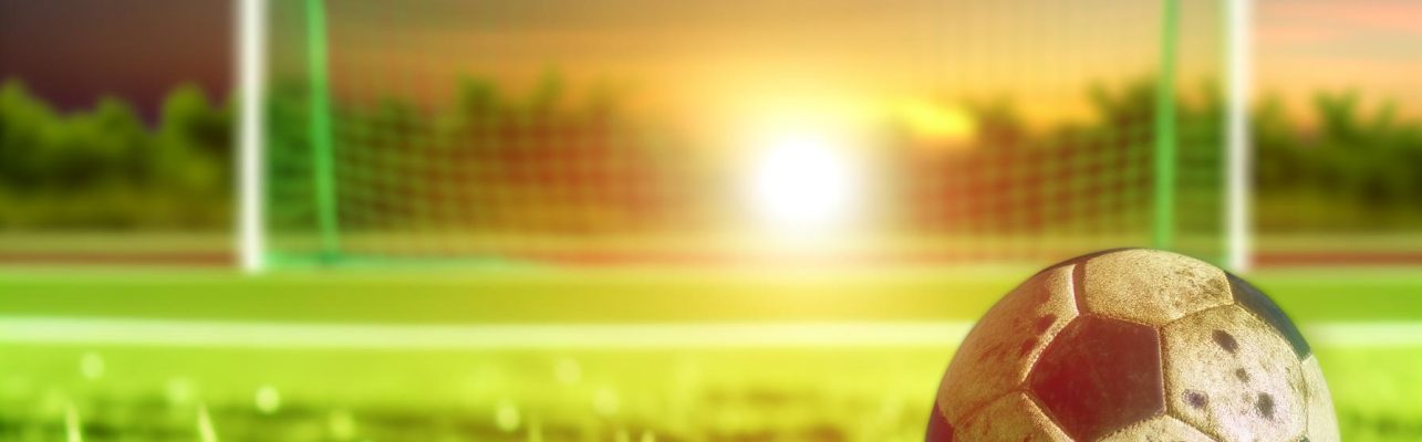 Un ballon de soccer use sur le gazon vert avec des buts en fond flou au coucher du soleil.