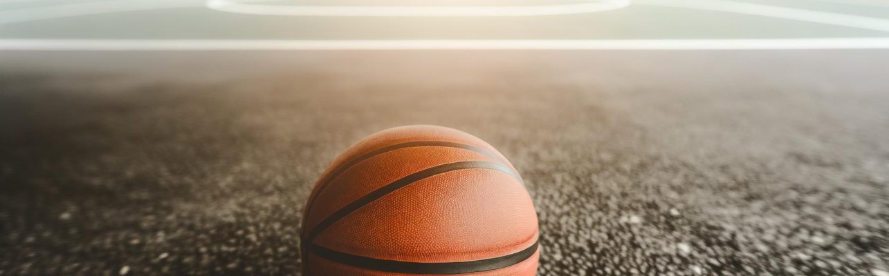 Un ballon de basketball repose sur un terrain exterieur en asphalte avec des arbres en fond flou.