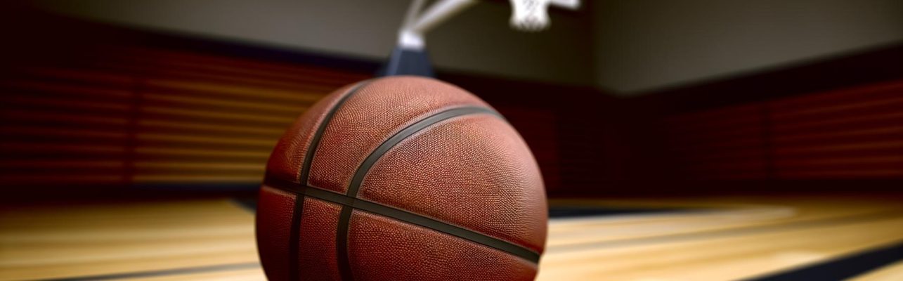 Un ballon de basketball repose sur le sol en bois d'un gymnase avec un panier flou en arriere-plan.