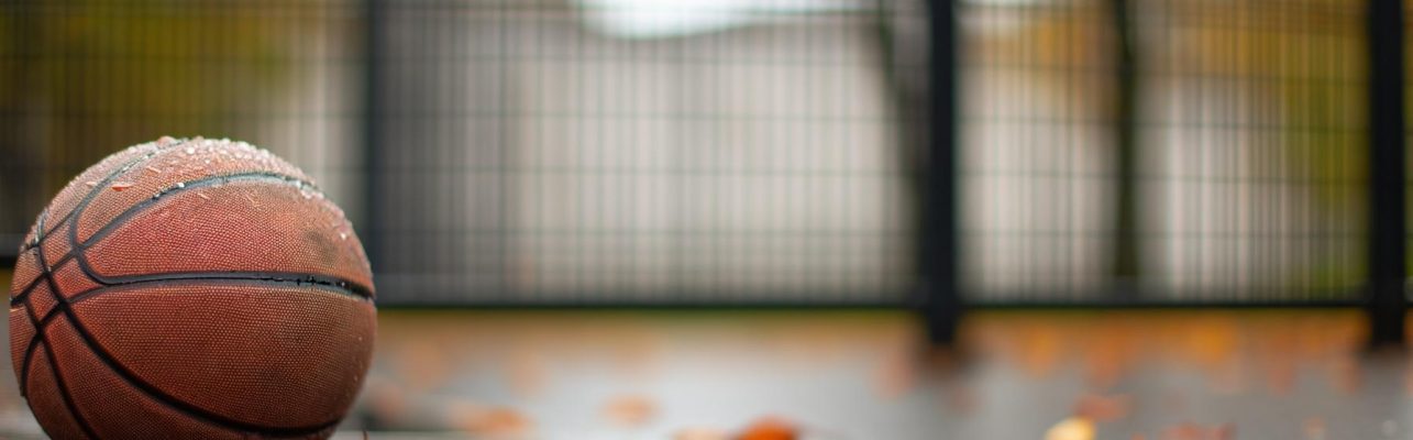Un ballon de basket repose sur un terrain extérieur humide avec des feuilles d'automne et un grillage en arrière-plan.