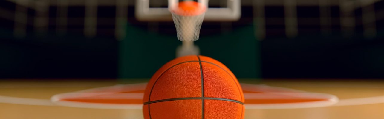 Un ballon de basket orange repose sur le parquet d'un terrain avec le panier flou à l'arrière-plan.