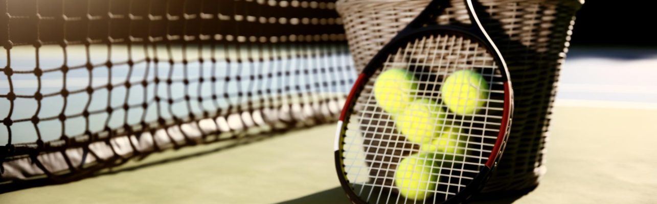 Raquette de tennis posee contre un panier de balles sur un court de tennis ensoleille.