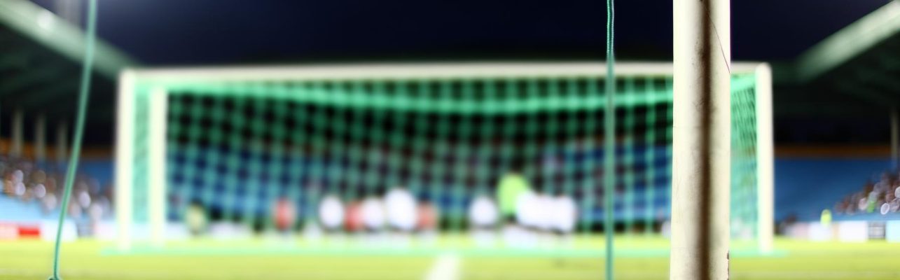 Stade de football lors d'un match en début de soirée avec un but inoccupé et des joueurs flous en mouvement.