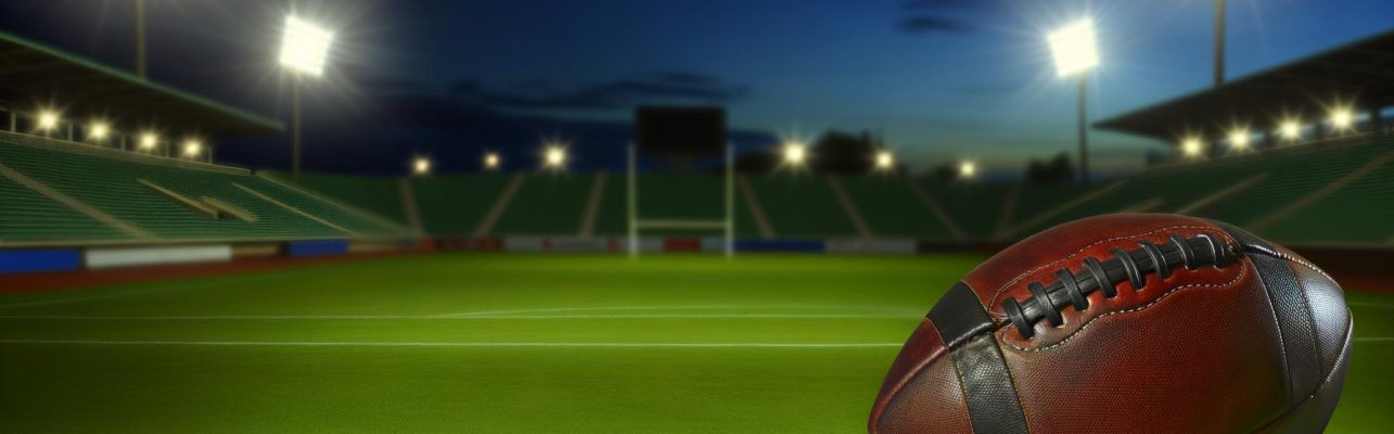 Un ballon de football en cuir est posé sur la pelouse d'un terrain de football vide au crépuscule, avec les lumières du stade qui s'allument.