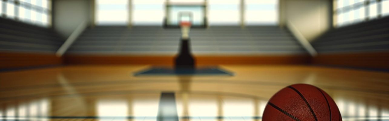 Un terrain de basketball vide avec un ballon posé sur le sol en bois dans un gymnase éclairé par la lumière naturelle.