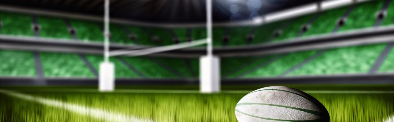 Un terrain de rugby avec un ballon posé sur la pelouse, les gradins et les poteaux visibles au loin.