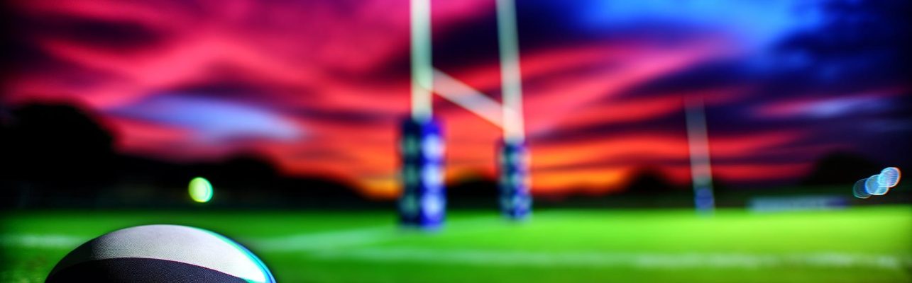 Un terrain de rugby au crépuscule avec des poteaux de but silhouettés contre un ciel coloré et un ballon de rugby sur l'herbe au premier plan.